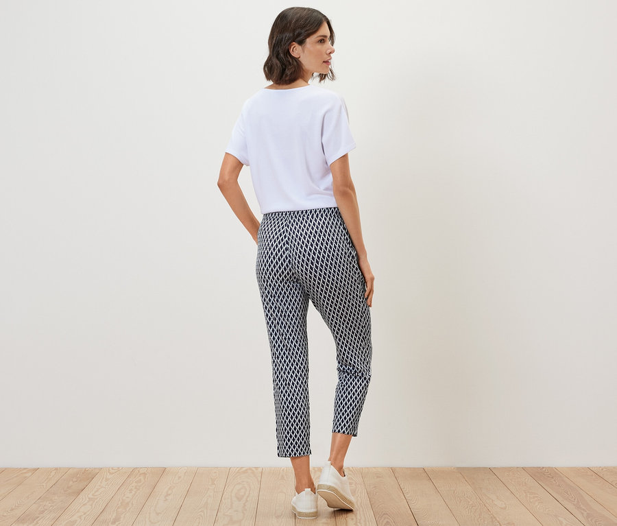 Žena stojí zády k fotoaparátu a má na sobě bílé tričko a vzorované žerzejové kalhoty.