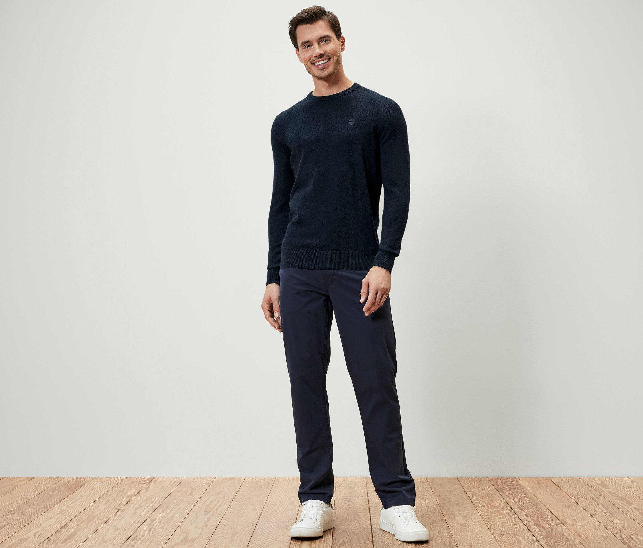 Muž stojí a má na sobě tmavě modrý svetr s kulatým výstřihem, tmavě modré kalhoty a bílé tenisky.