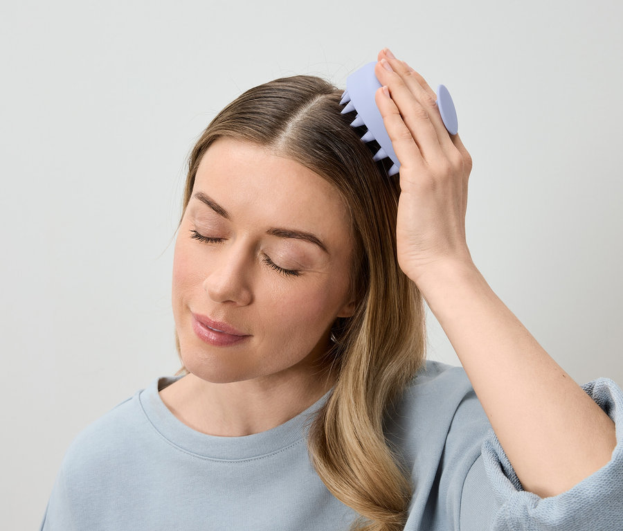 Žena si masíruje hlavu světle fialovým masážním kartáčem na vlasy.