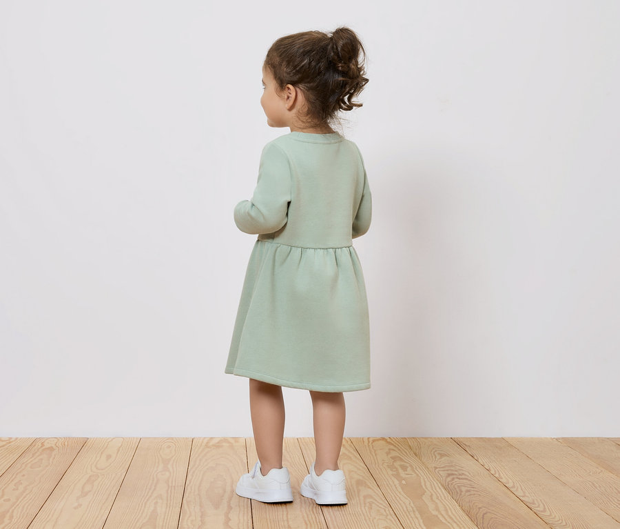 Holčička v zelených šatech a bílých botách stojí zády ke kameře.