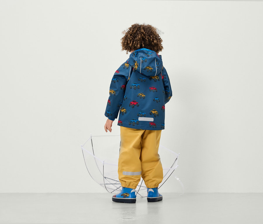 Chlapec v bundě s potiskem aut a žlutých kalhotách stojí zády k fotoaparátu vedle průhledného deštníku.