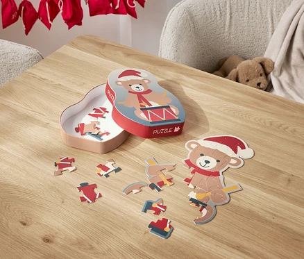 Na stole leží rozložené puzzle s motivem medvídka v santovské čepici a jeho krabička.