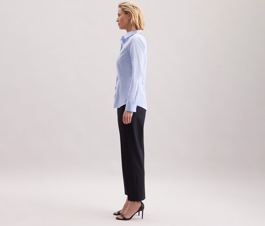 Žena stojí bokem a má na sobě světle modrou halenku Seidensticker Slim Fit non iron.