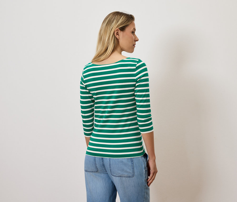 Žena s blond vlasy stojí zády ke kameře a má na sobě tričko s tříčtvrtečním rukávem se zelenými a bílými pruhy a modré džíny.