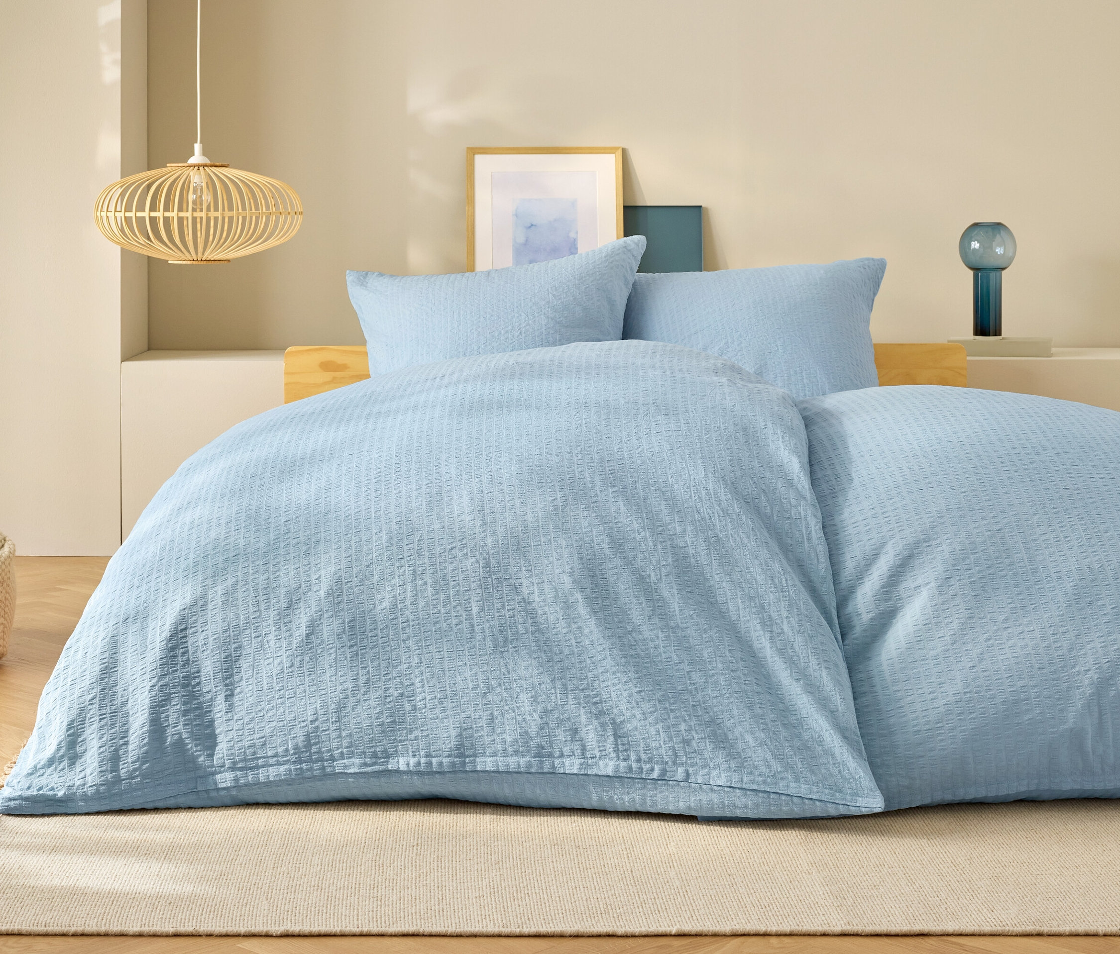 Světle modré ložní prádlo na posteli s polštáři a lampou v pozadí.