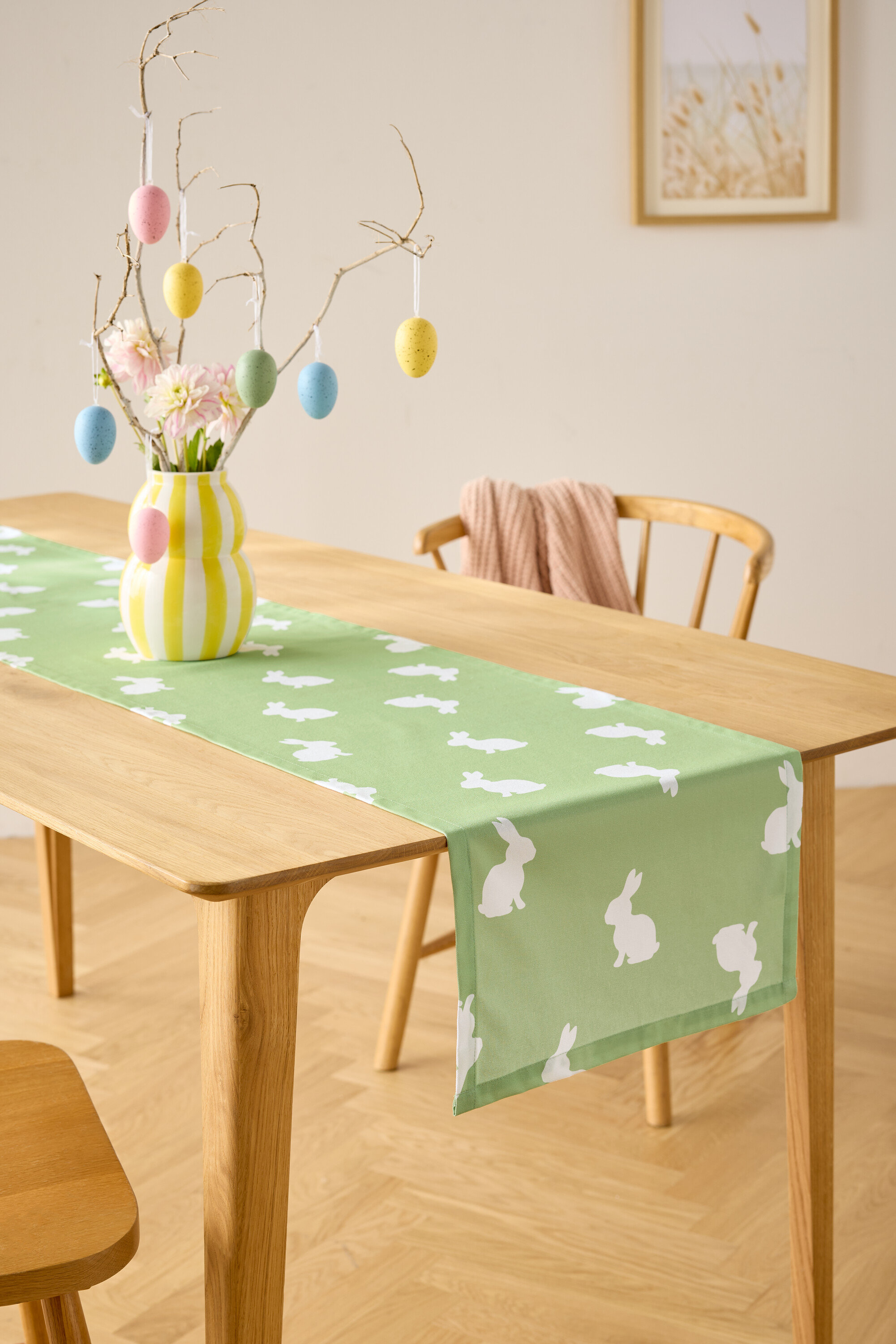 Jídelní stůl se světle zeleným běhounem s bílými motivy zajíčků, váza s květinami a dekorace s velikonočními vajíčky.