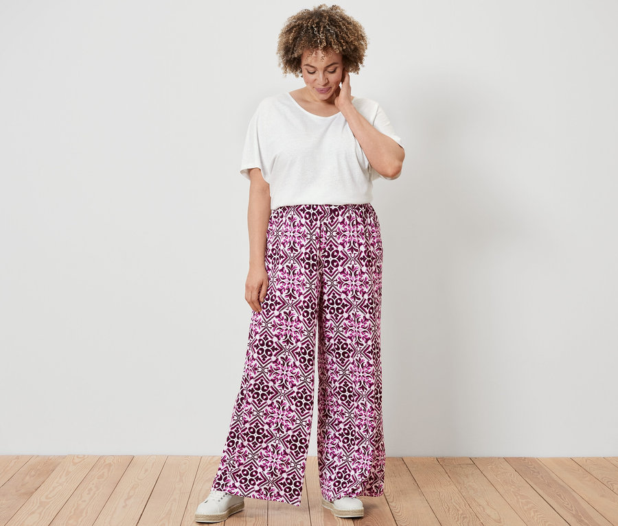 Žena v bílém tričku ze směsi bavlny a lnu a v bobulových kalhotách se širokými nohavicemi s potiskem stojí na dřevěné podlaze.