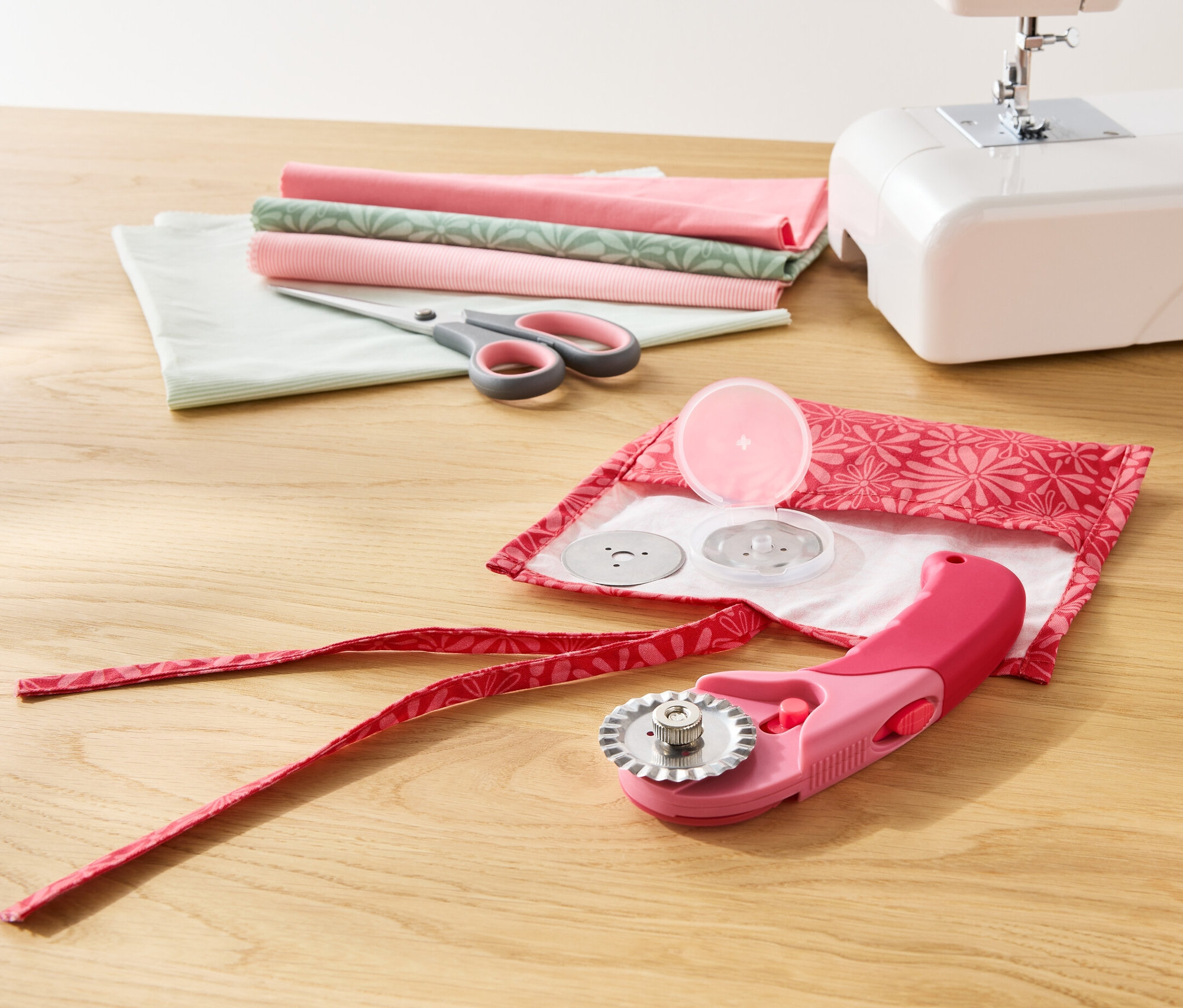 Na dřevěném stole leží bílý šicí stroj, nůžky, látky a látkový rotační ořezávač.