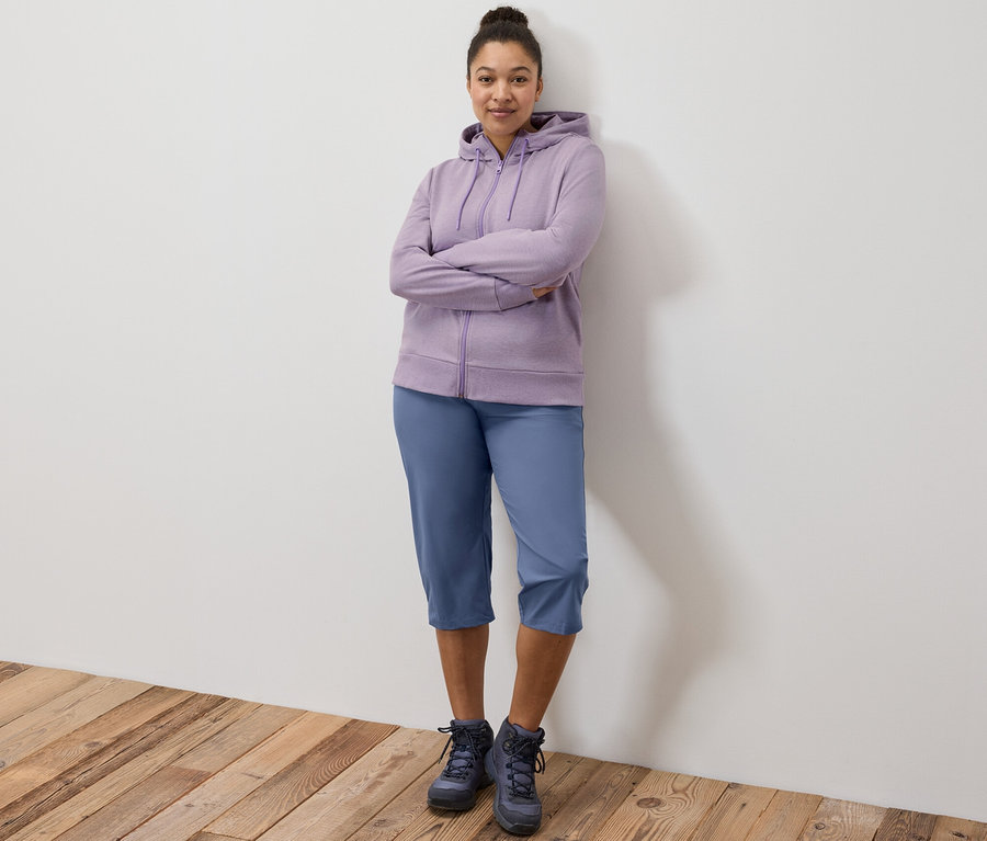 Žena stojí s rukama zkříženýma na prsou, má na sobě fialovou mikinu a funkční capri kalhoty.