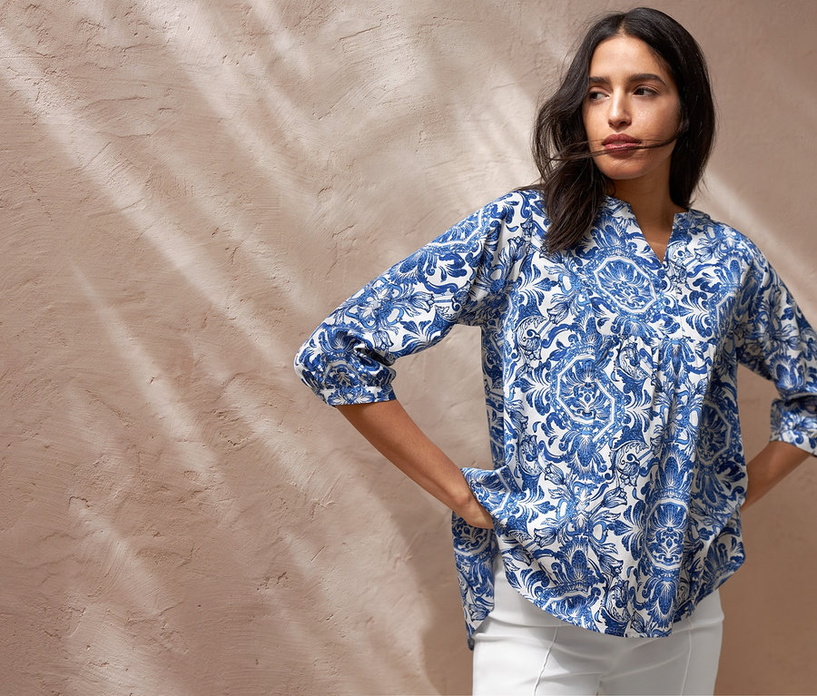 Žena pózuje v tunikové halence s potiskem, bílých strečových kalhotách v 7/8 délce a teniskách. Má tašku přes rameno.