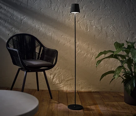 Černá LED stojací lampa na dobíjení pro venkovní použití stojí vedle černého proutěného křesla a rostliny v květináči.