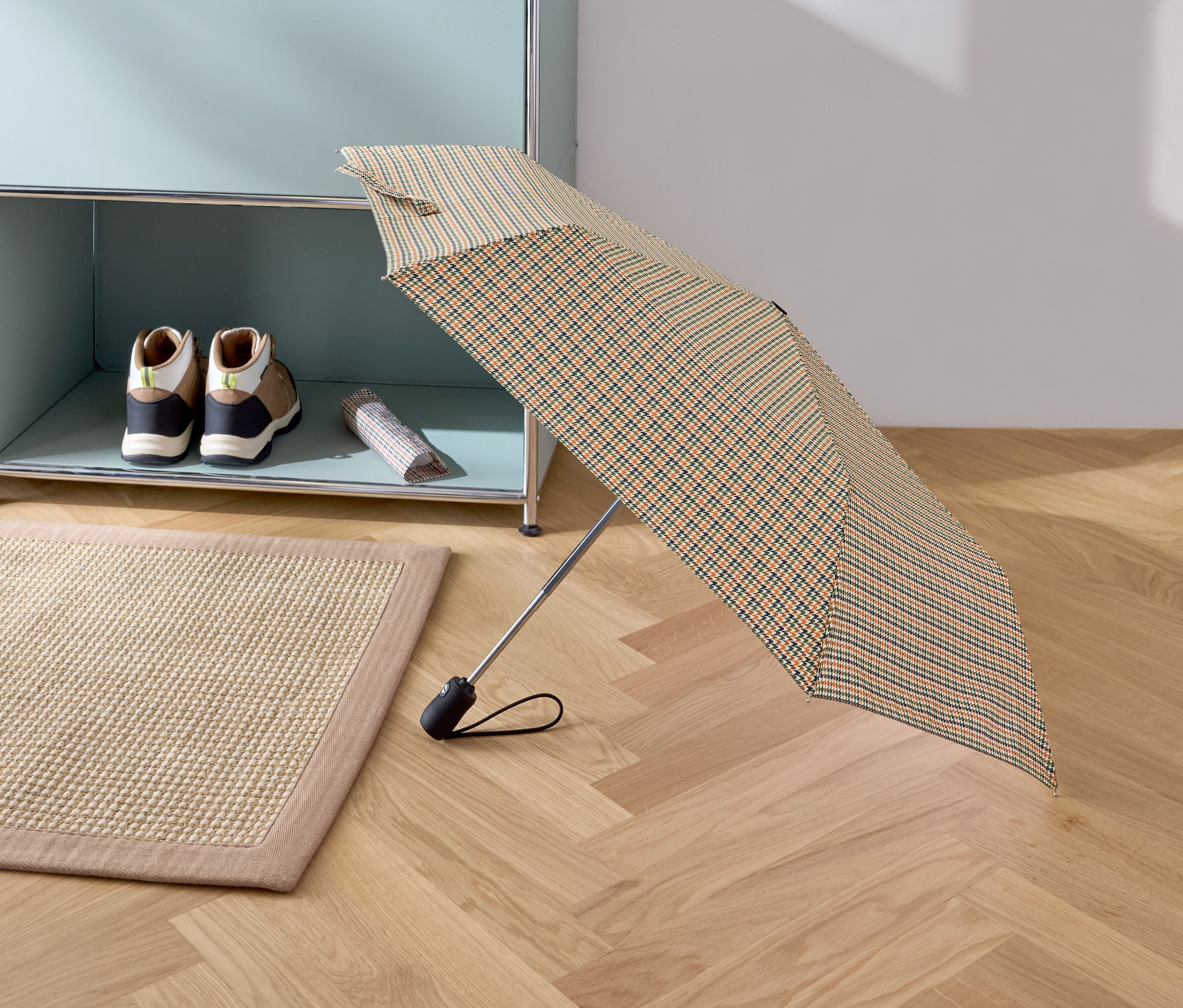 Otevřený skládací deštník s kostkovaným vzorem stojí na parketové podlaze. V pozadí je béžový koberec, boty a taška na poličce.