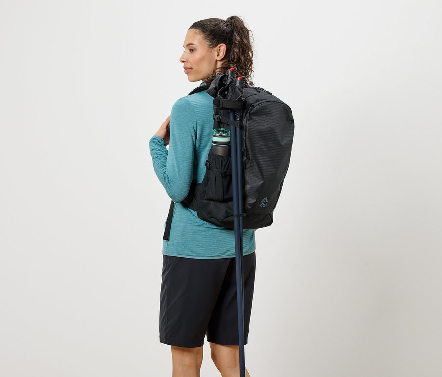 Žena zezadu s černým batohem, lahví na pití a trekovými holemi, oblečená v tyrkysovém topu a černých šortkách.