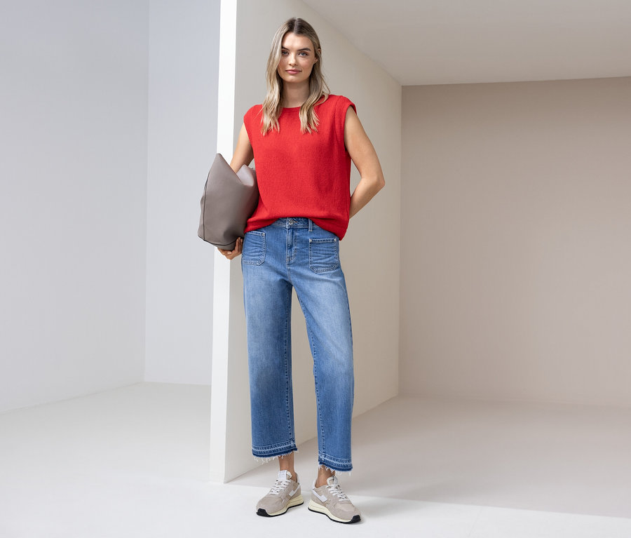 Žena v červeném topu, džínách a teniskách stojí se šedou taškou ve světlé místnosti.