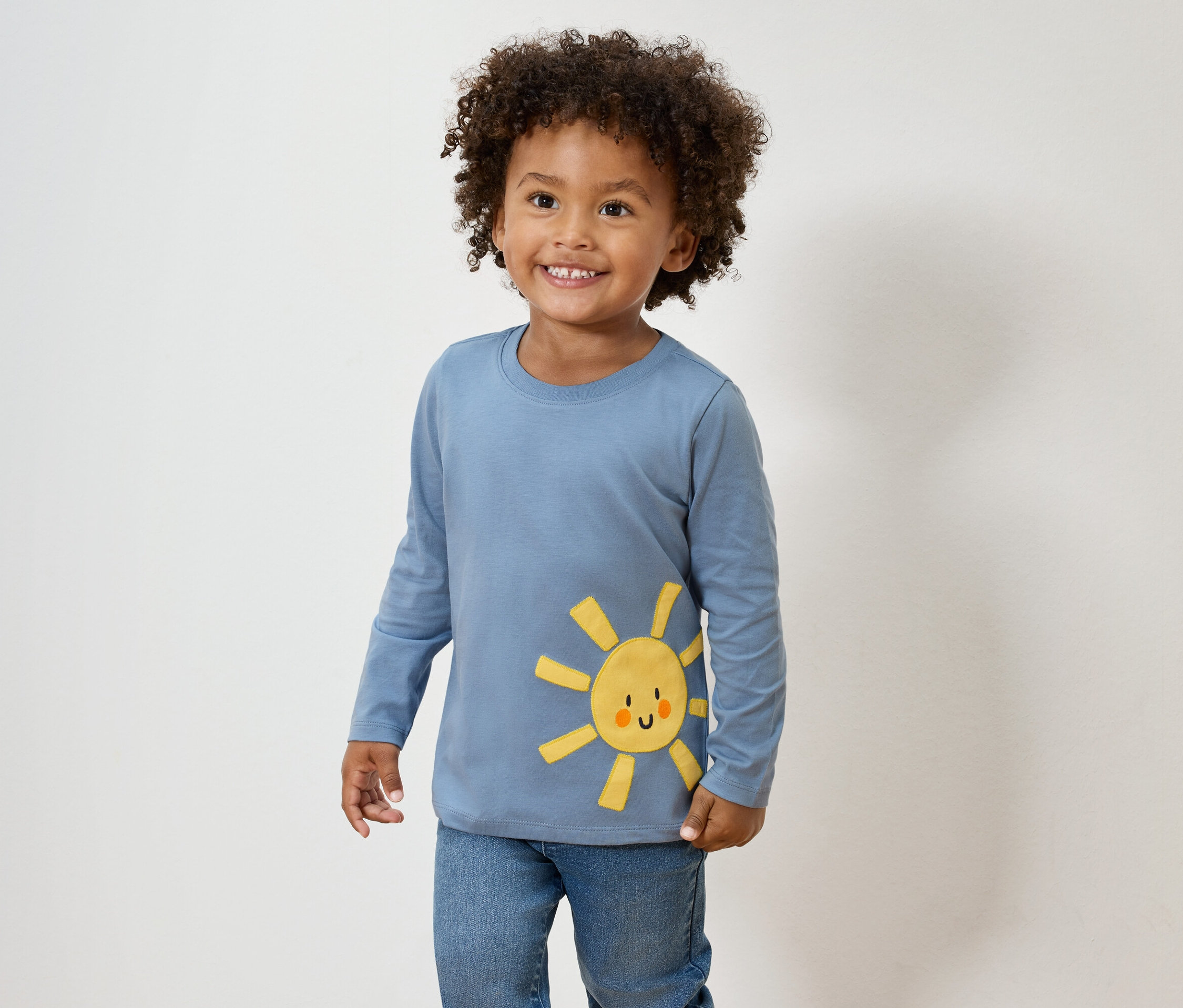 Usmívající se chlapec s kudrnatými vlasy má na sobě modré tričko s dlouhým rukávem se žlutou aplikací slunce a džíny.