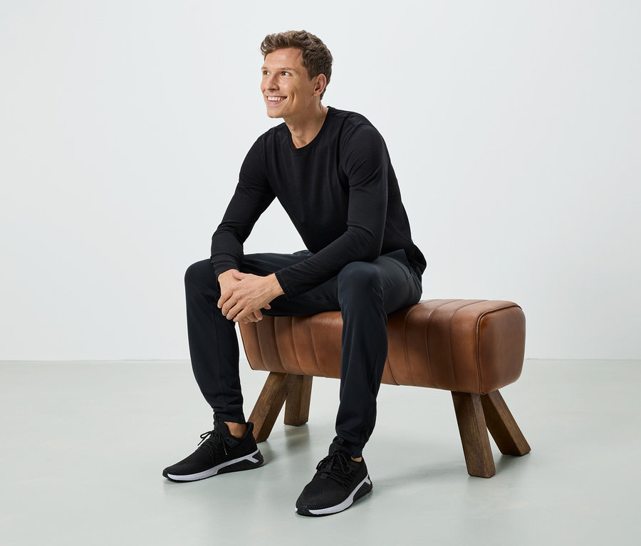 Muž sedí na hnědé lavici. Má na sobě černé tričko s dlouhým rukávem, černé kalhoty a černé tenisky.