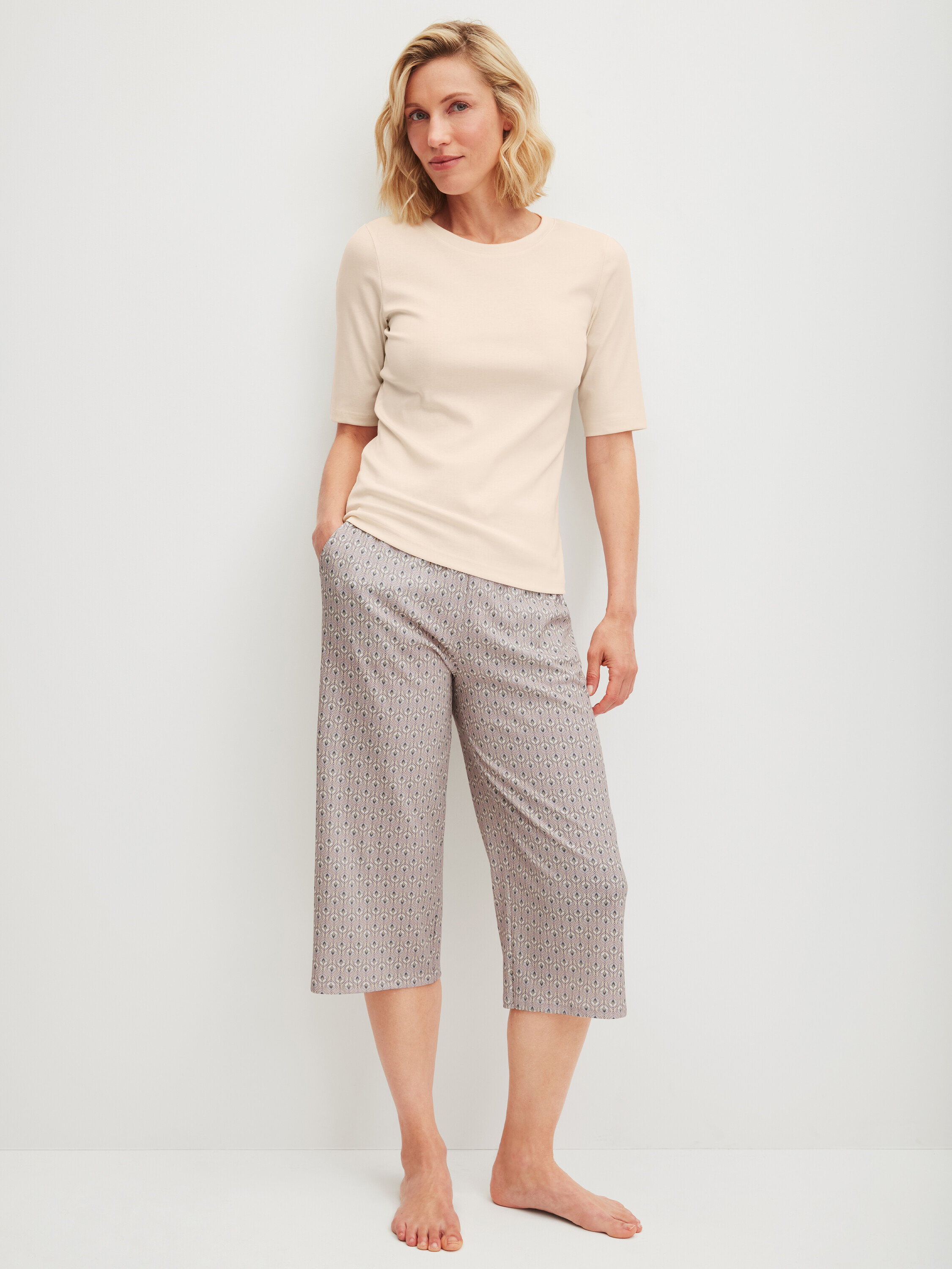 Žena stojí bosá, má na sobě světlé tričko a vzorované tříčtvrteční kalhoty.