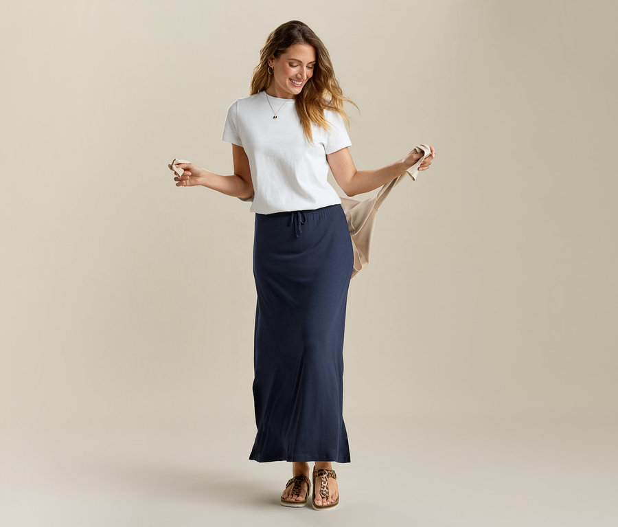 Žena drží béžový kardigan, má na sobě bílé tričko a dlouhou džínovou sukni v tmavě modré barvě.