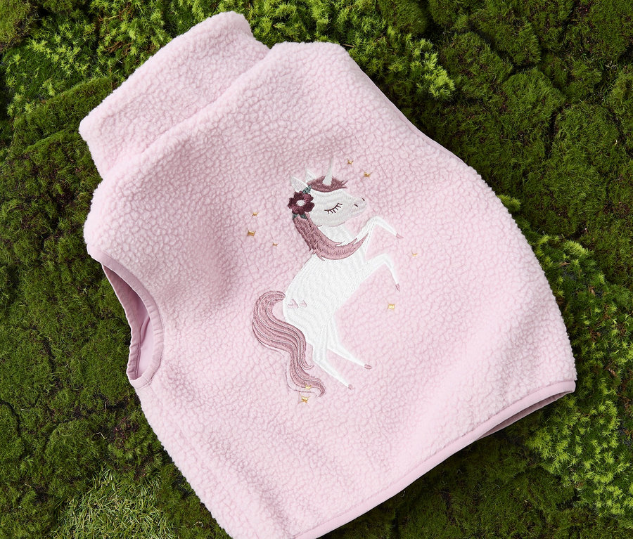 Růžová fleecová vesta s aplikací jednorožce leží na zeleném mechu.