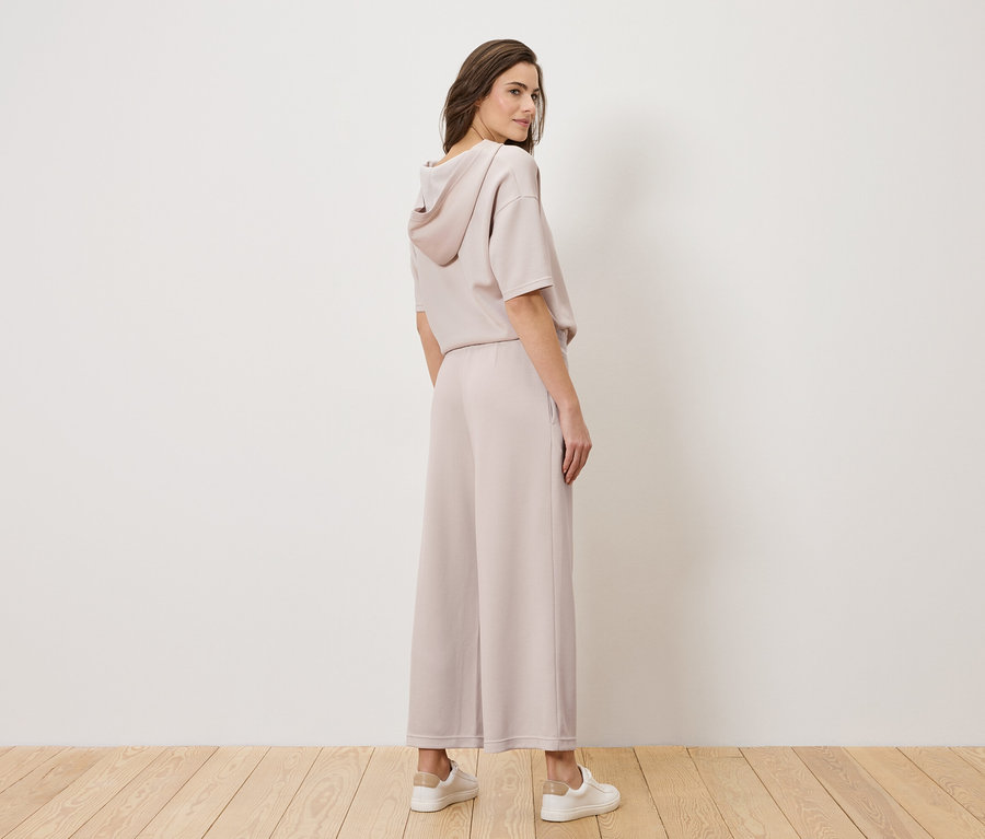 Žena stojí ve světle růžovém kalhotovém kostýmu a bílých teniskách, otáčí hlavu přes rameno.