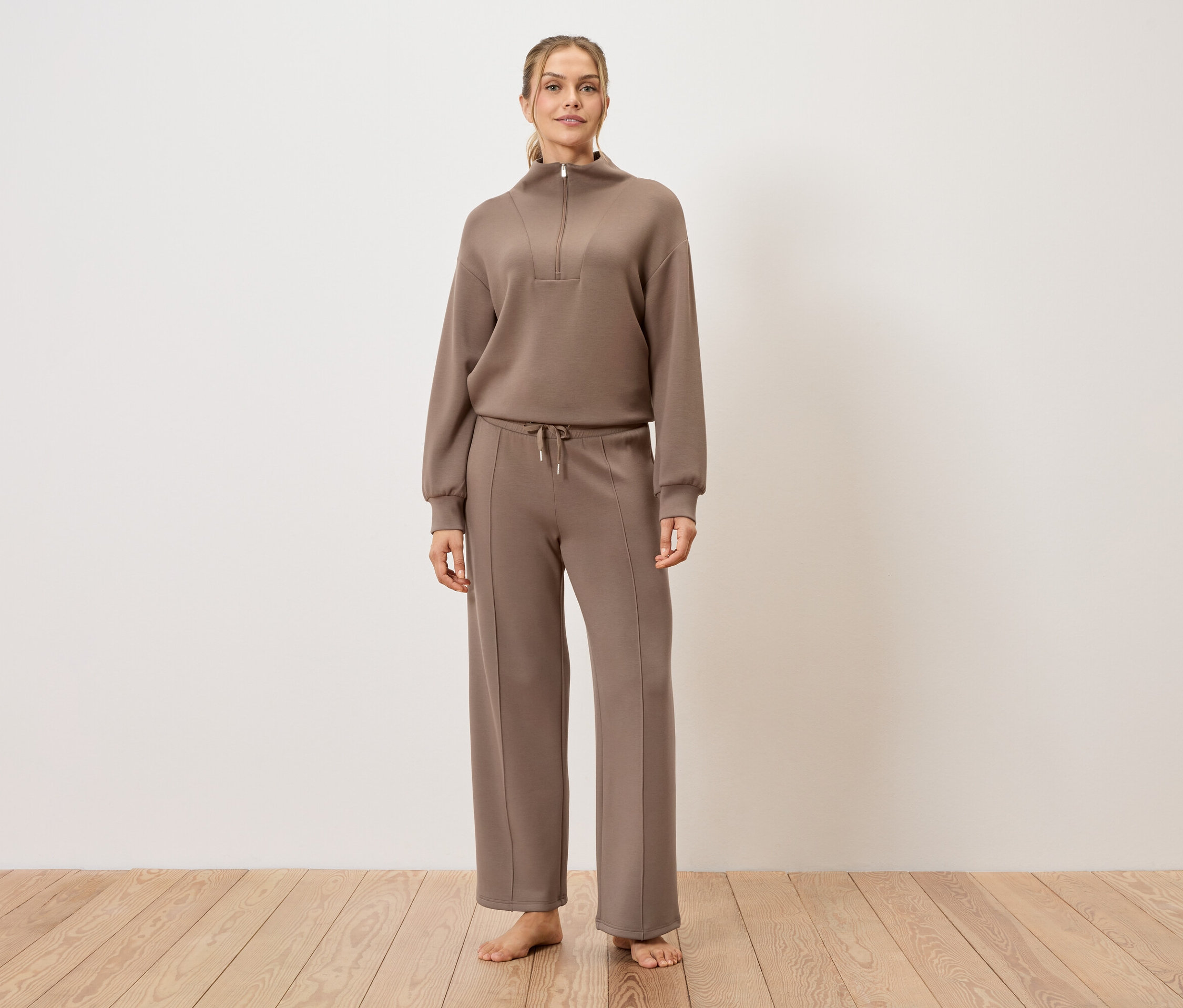 Žena stojí bosá na dřevěné podlaze a má na sobě hnědou teplákovou soupravu.