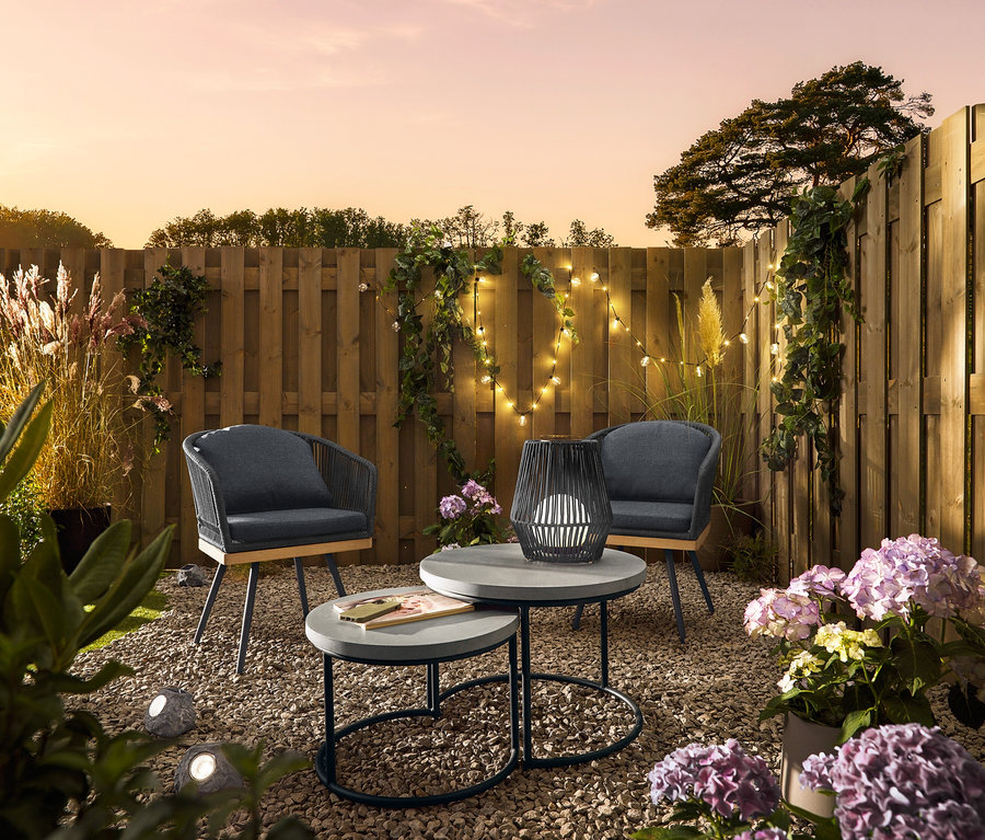 Dvě otočné zahradní židle s textilním křížovým výpletem stojí vedle 2 zahradních konferenčních stolků s polycementovou deskou. Na stole stojí malá solární lucerna s textilní šňůrkou. Prémiové solární LED kameny leží ve štěrku.