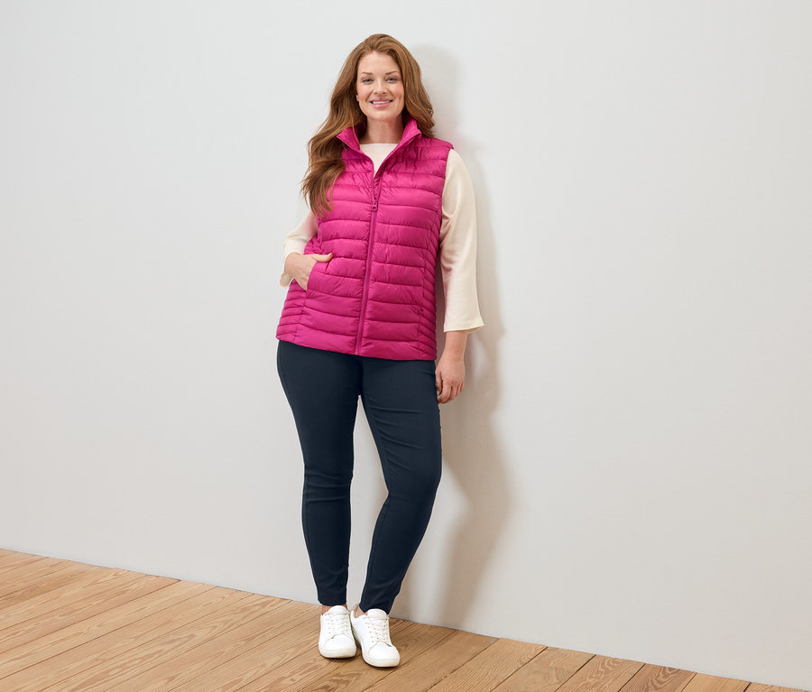 Žena v růžové prošívané vestě, béžové halence, modrých džínách a bílých teniskách stojí u bílé zdi.