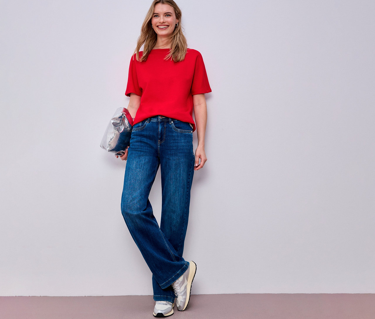 Žena v červeném tričku s krátkým rukávem Street One, modrých džínách a stříbrných teniskách drží stříbrnou tašku.