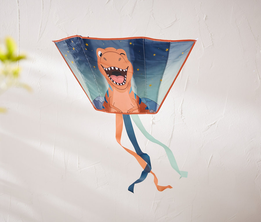 Létající drak ve tvaru lichoběžníku s usmívajícím se oranžovým dinosaurem a hvězdami visí na texturované bílé zdi. Dole visí oranžové, tmavě modré a světle modré stuhy.