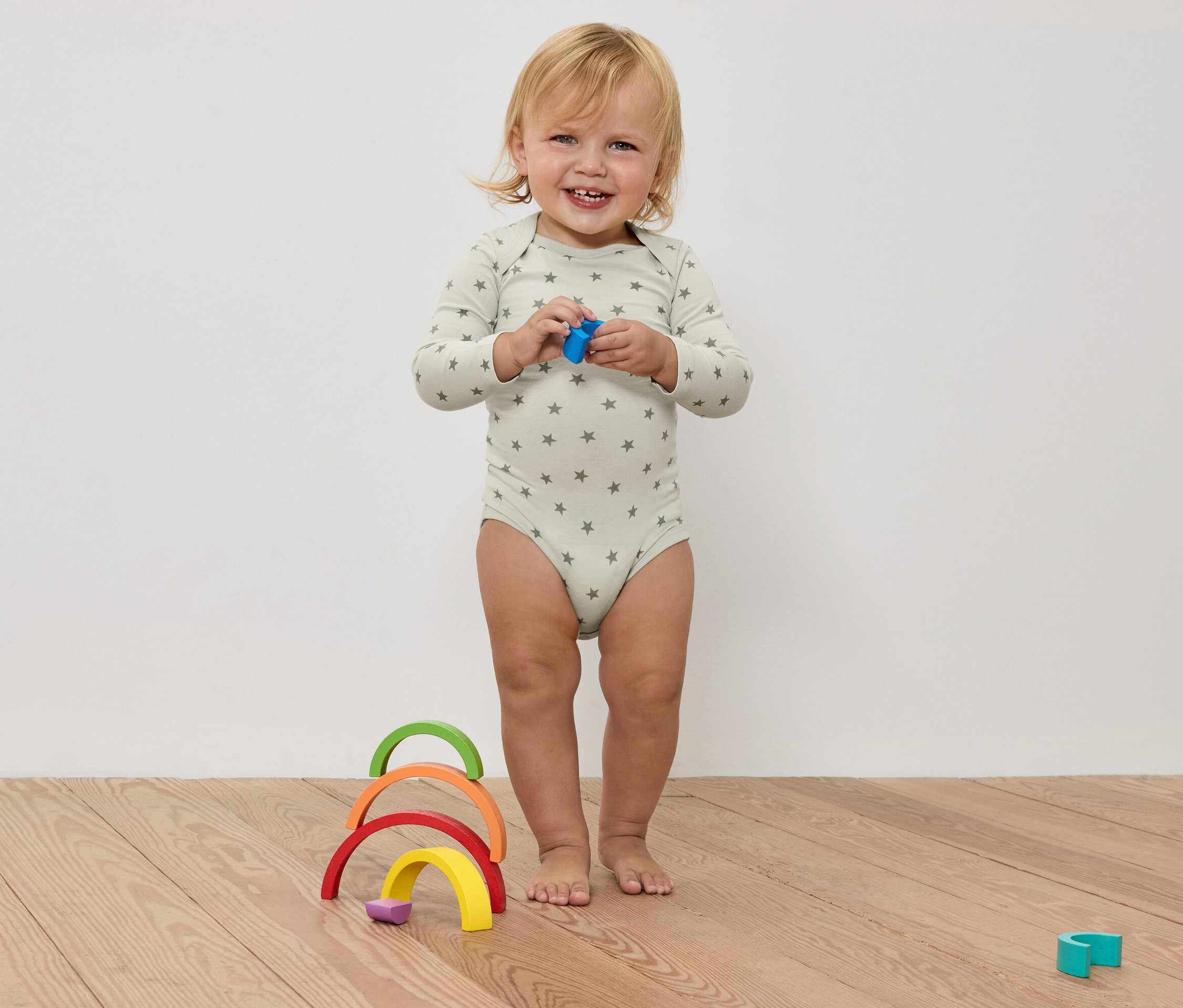 Usmívající se batole v body s hvězdami drží hračku v rukou, před barevnou dřevěnou duhou na dřevěné podlaze.