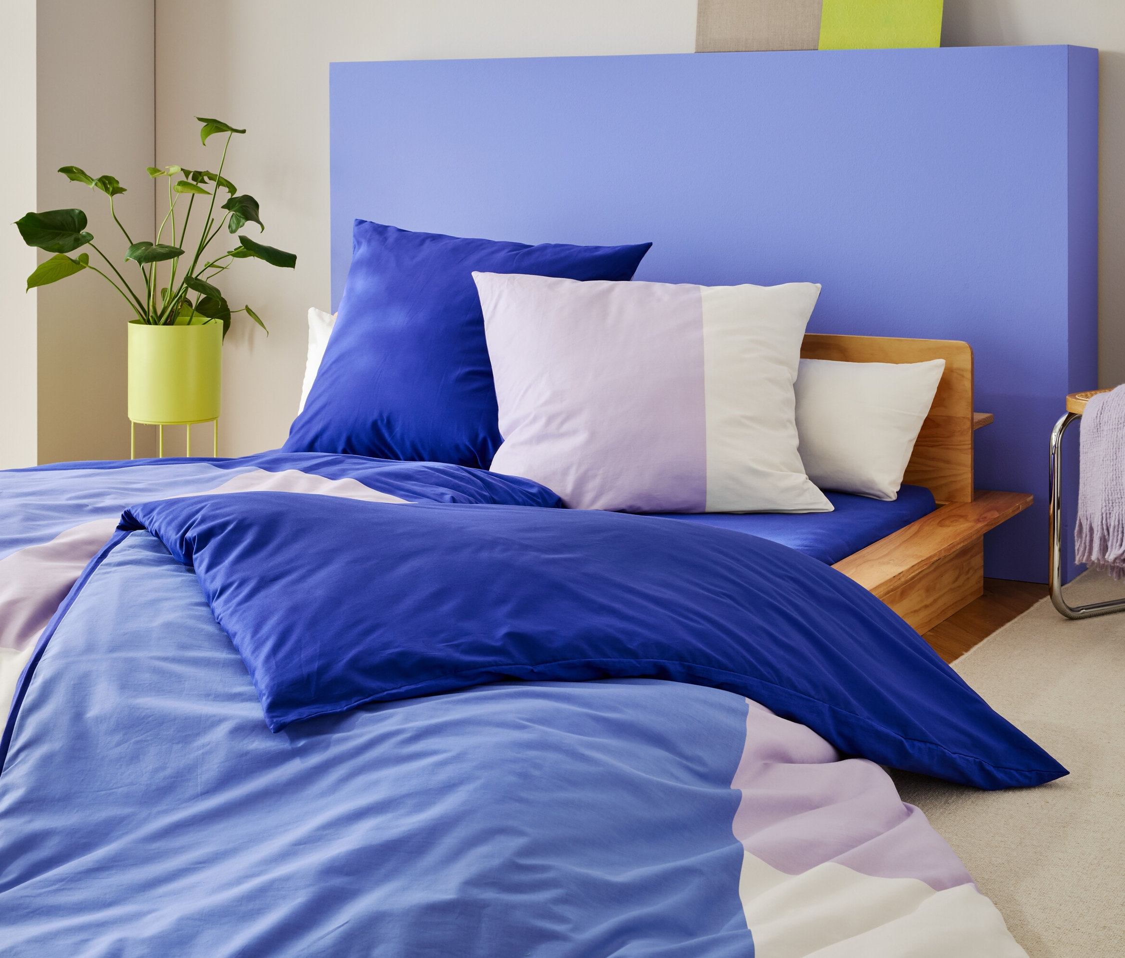 Postel s modro-fialovým ložním prádlem a polštáři stojí před modrou stěnou, vedle ní rostlina a židle s dekou.