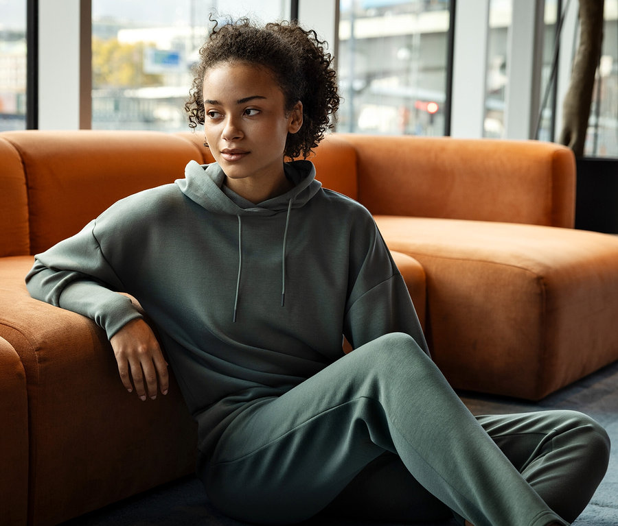 Žena sedí na zemi, opírá se o oranžovou pohovku a má na sobě zelenou mikinu s kapucí a tepláky.