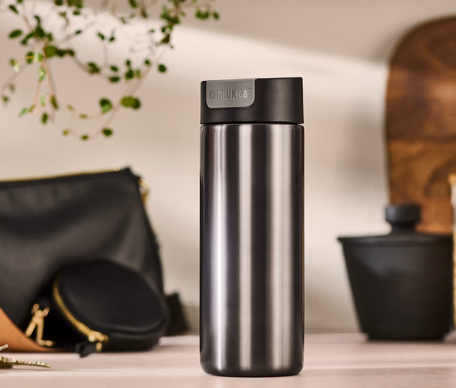 Šedý cestovní hrnek Kambukka o objemu 500 ml stojí na stole vedle kabelky, pouzdra na mince a klíčů.