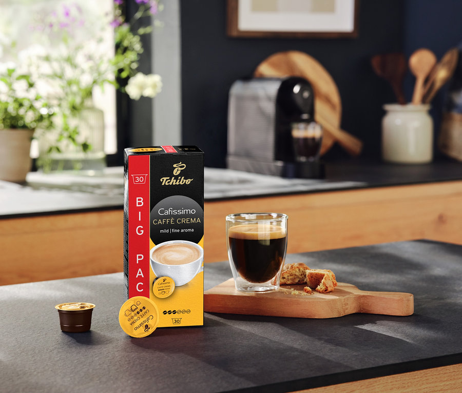 Balení kapslí Cafissimo Caffè Crema mild, šálek kávy a kávovar na kuchyňské lince.
