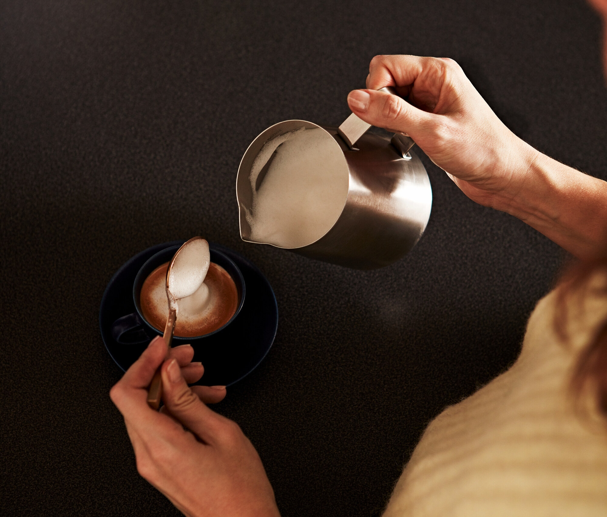 Žena nalévá napěněné mléko z malé nerezové konvičky do šálku s kávou, lžičkou nabírá pěnu.