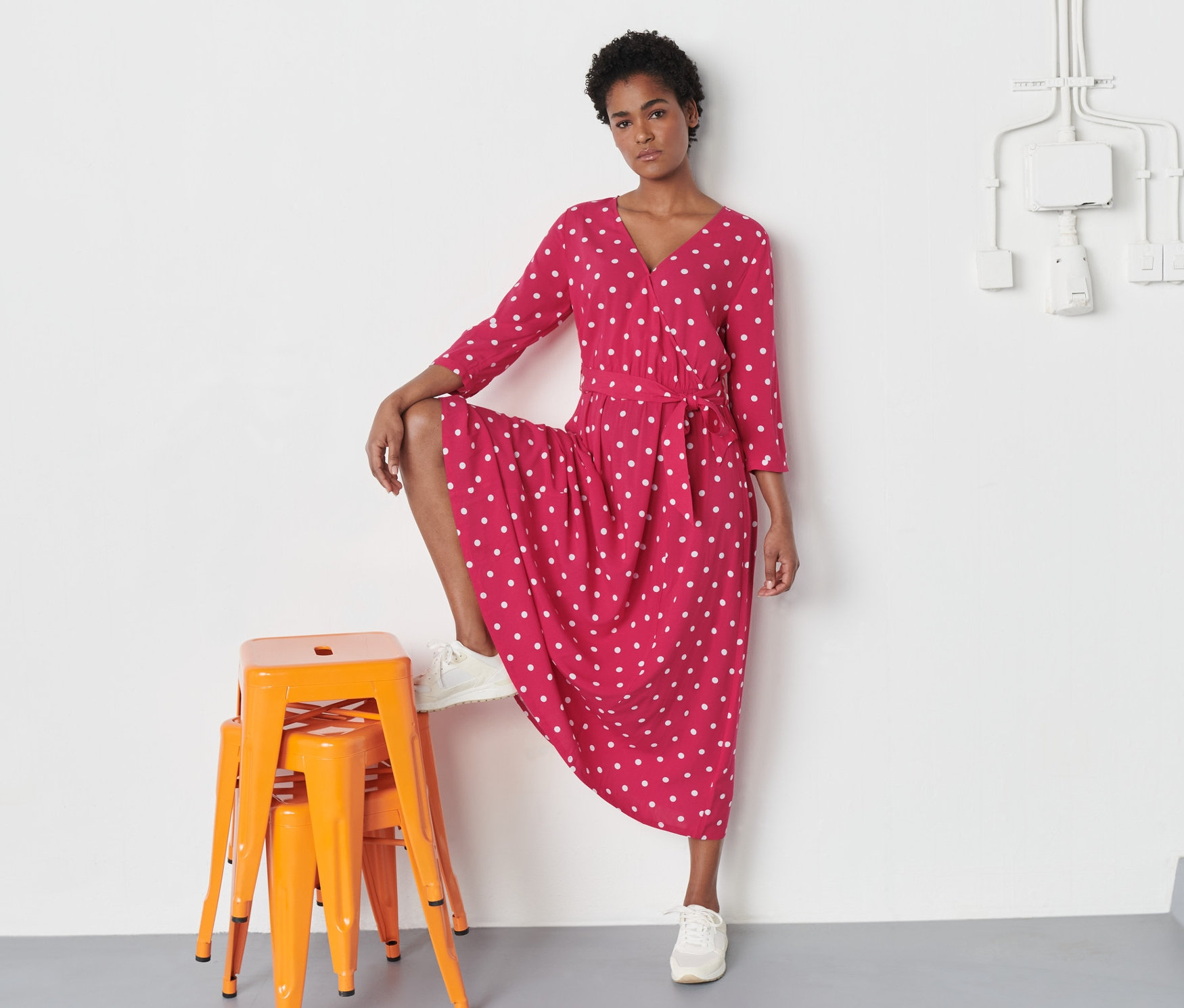 Žena pózuje v puntíkovaných šatech v zavinovacím vzhledu a bílých teniskách, nohu má na oranžové stoličce.