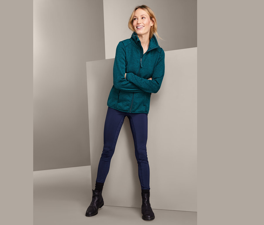 Žena pózuje v bundě z pleteného fleecu s recyklovaným materiálem, tmavě modrých jezdeckých kalhotách a černých botách.