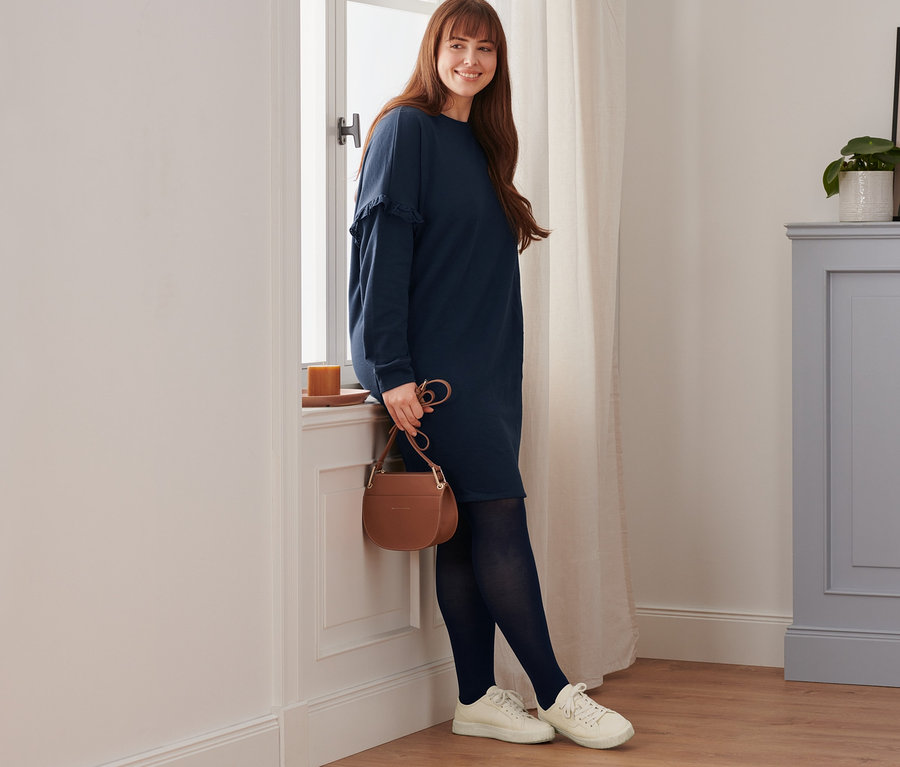 Žena se usmívá a opírá se o okno. Má na sobě teplákové šaty, kožené boty sneaker, neprůhledné punčochové kalhoty (2 ks) a kabelku.