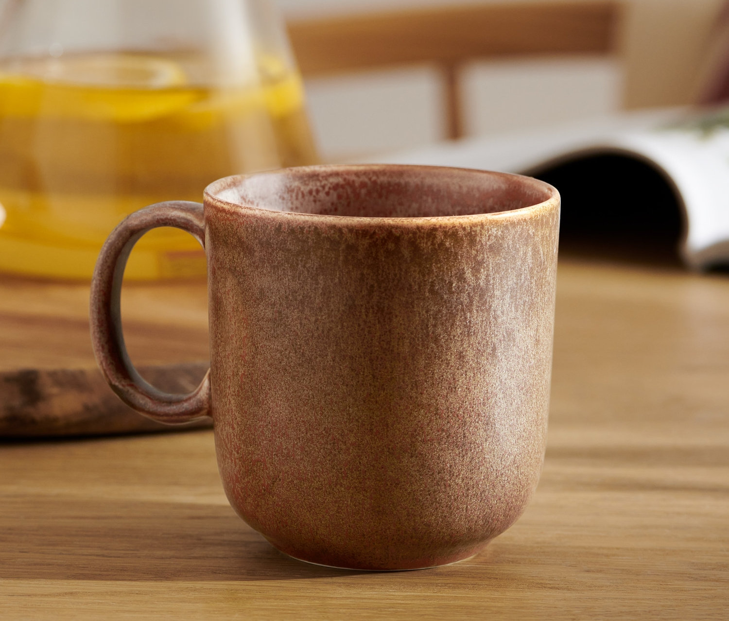 Hnědý hrnek na stole s nápojem a sušenkami v pozadí.