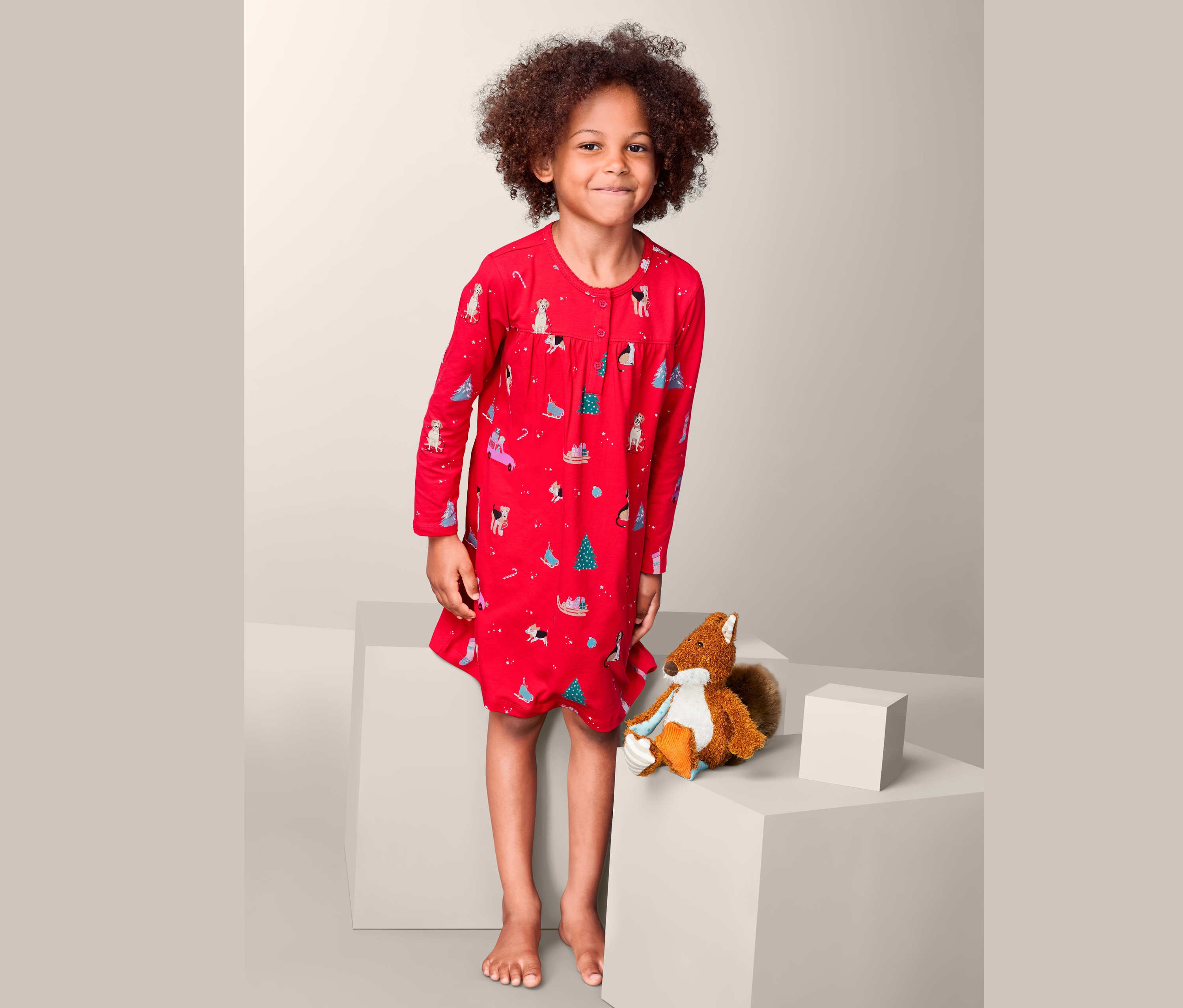 Dívka stojí a má na sobě červenou noční košili s potiskem psů a vánočních stromků. Vedle ní sedí plyšová veverka.