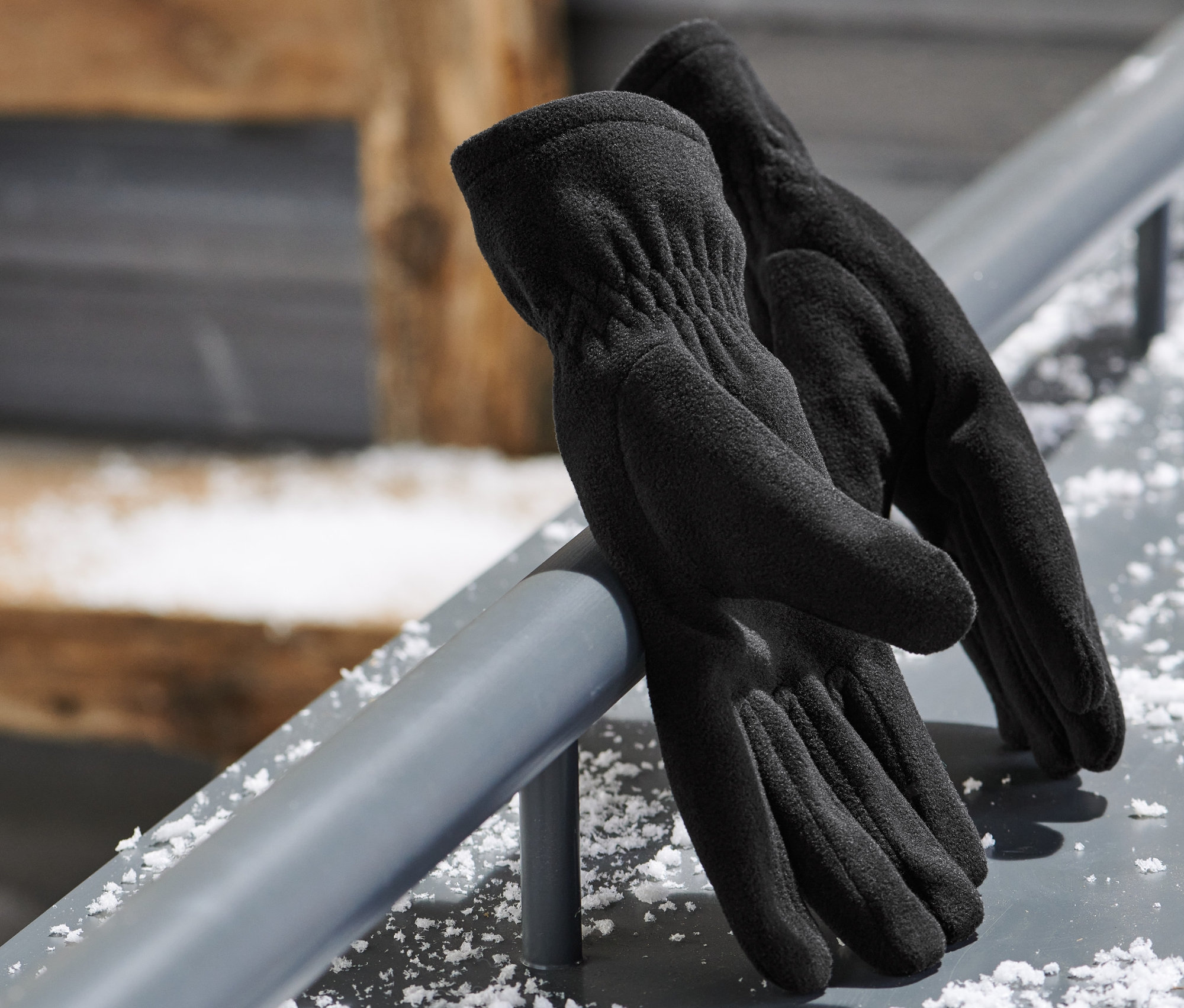Černé mikrofleecové rukavice leží na kovové tyči. V pozadí jsou lyžařské brýle.