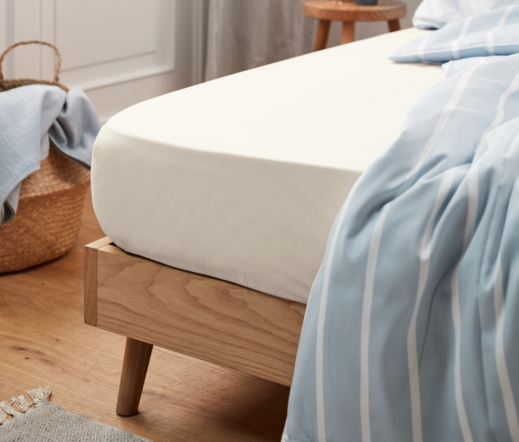 Béžové prostěradlo Jersey, standardní velikosti, natažené na posteli s dřevěným rámem. V pozadí je koš s modrou látkou.