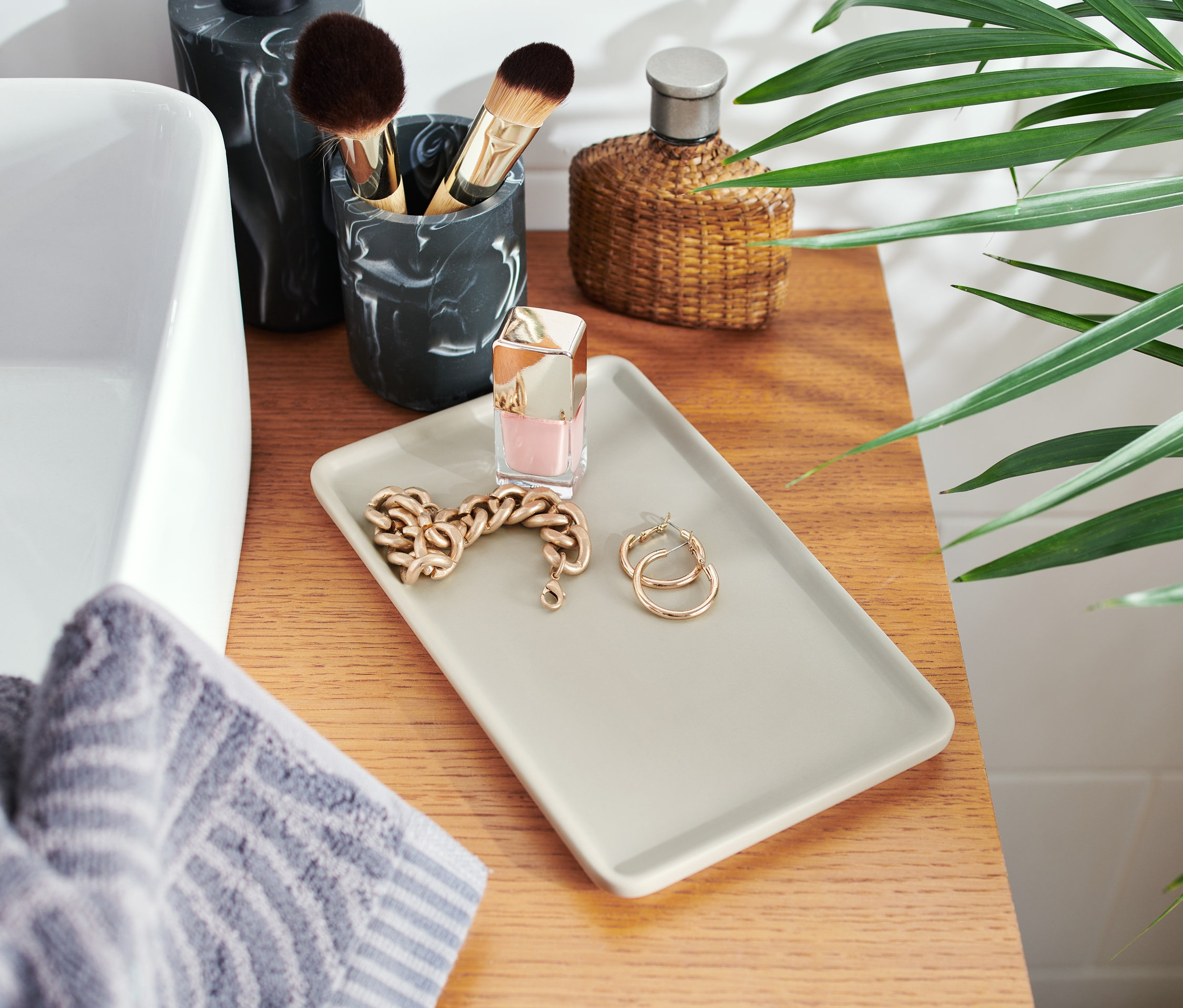 Keramická podložka s řetízkem, náušnicemi a lakem na nehty na dřevěné desce vedle umyvadla.