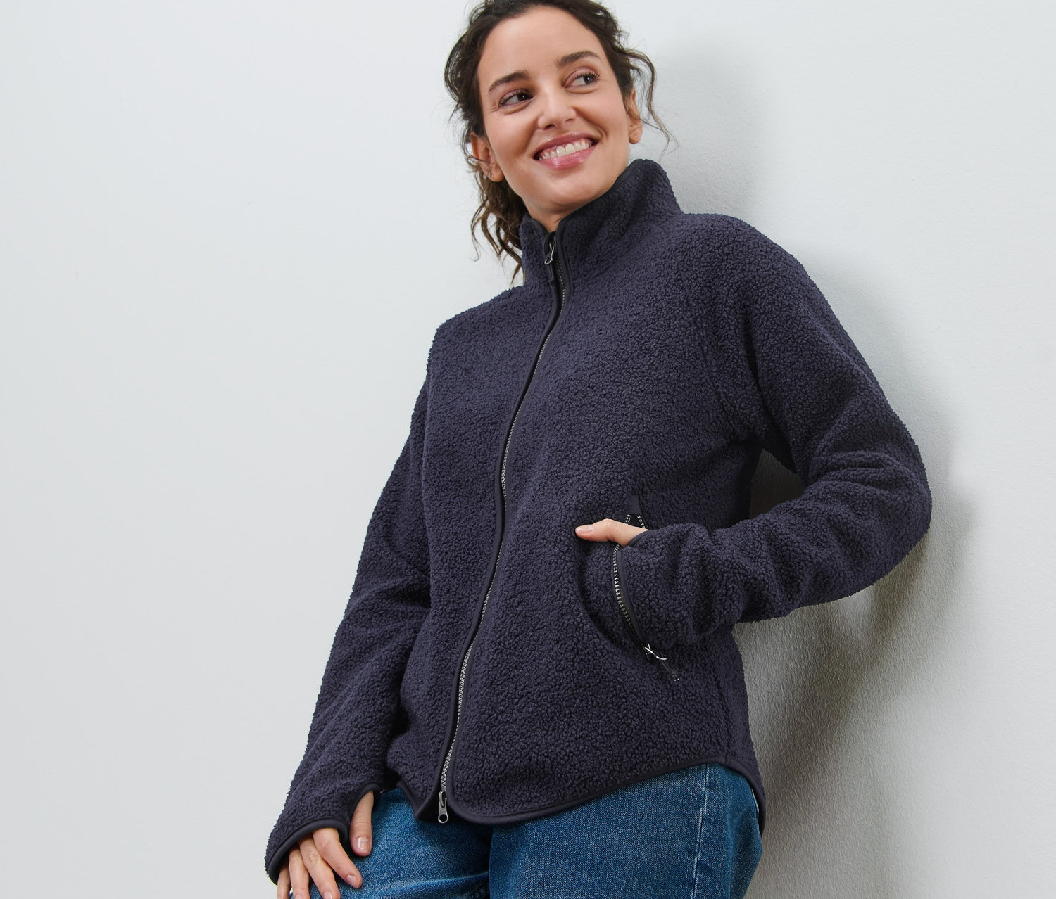 Usmívající se žena opřená o zeď má na sobě námořnickou modrou bundu z plyšového fleecu a džíny.