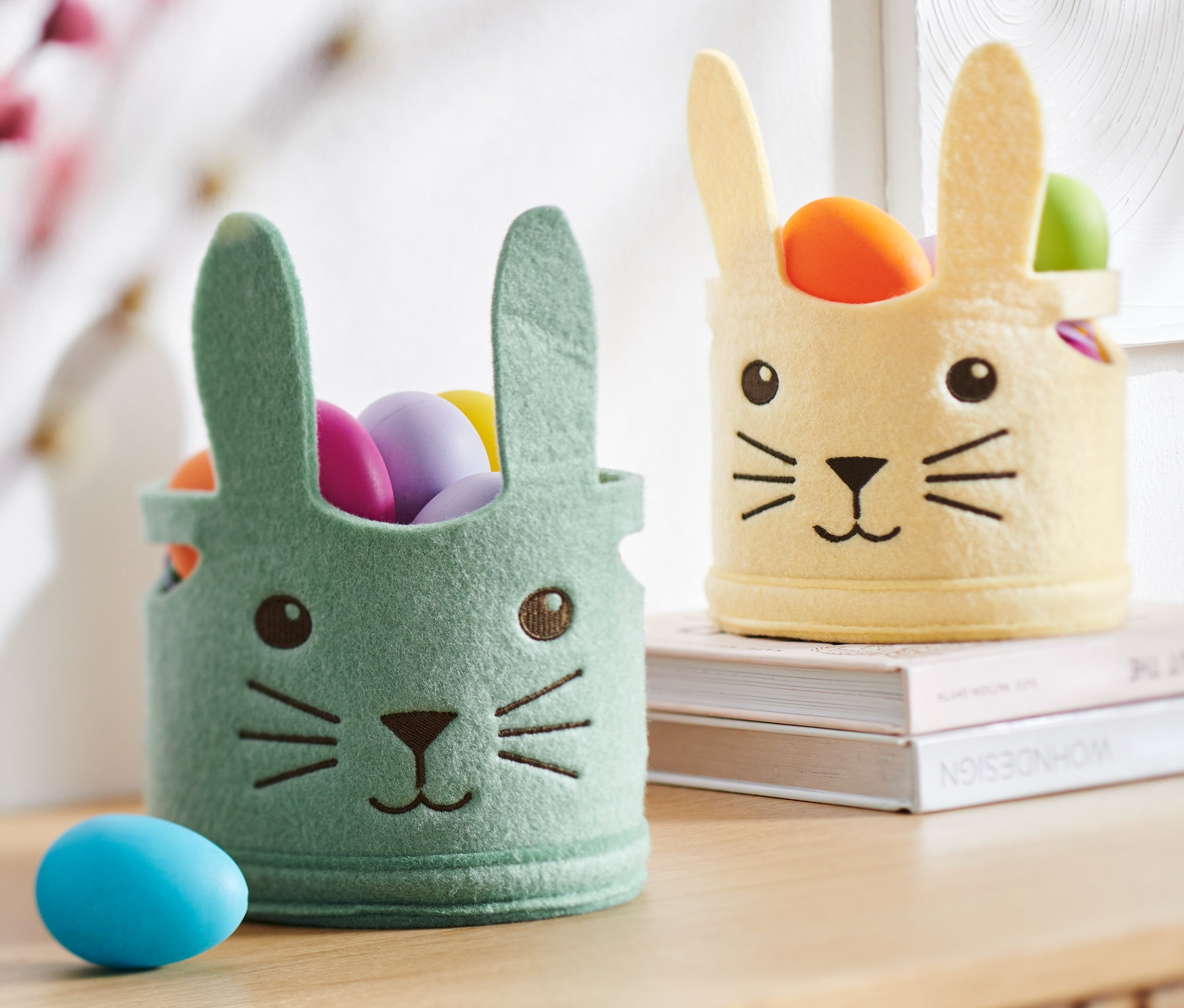 Dva velikonoční košíčky s obličejem králíka plné barevných vajíček stojí na skříňce.