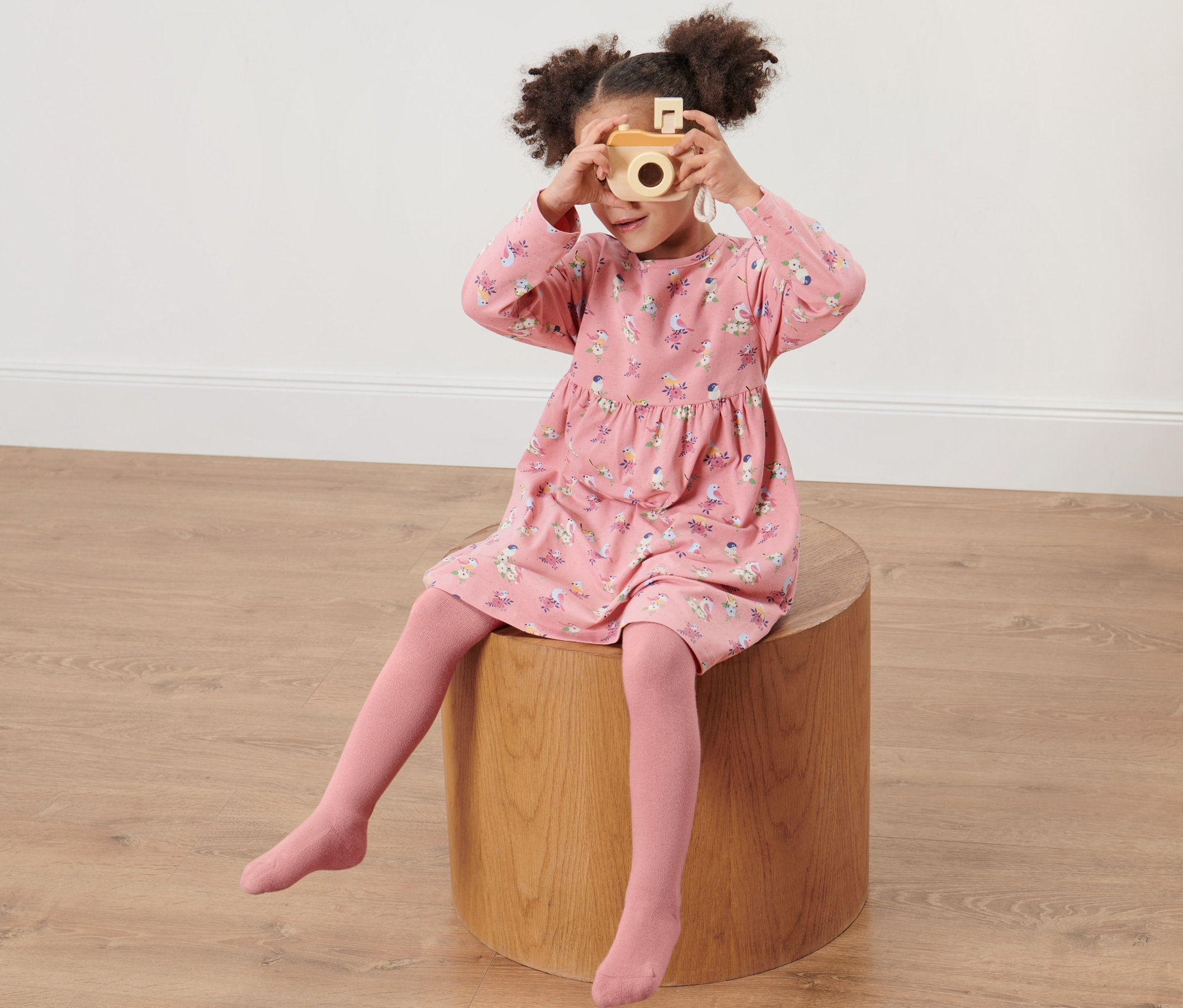 Dívka sedí na dřevěné stoličce a fotí dřevěným fotoaparátem. Má na sobě růžové šaty s potiskem a růžové dětské punčocháče, 2 ks.