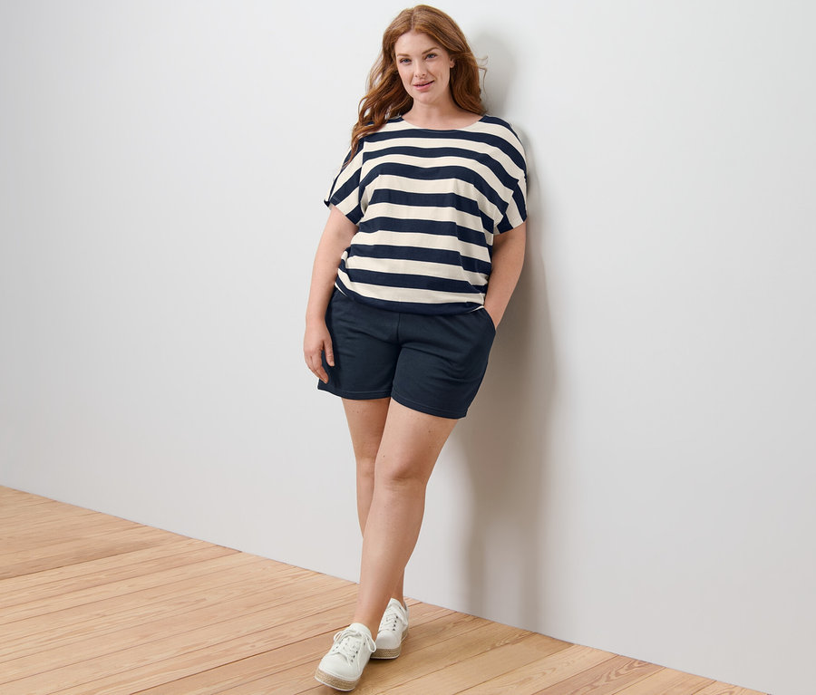 Žena stojí opřená o zeď a má na sobě halenkové tričko s proužky a teplákové šortky.