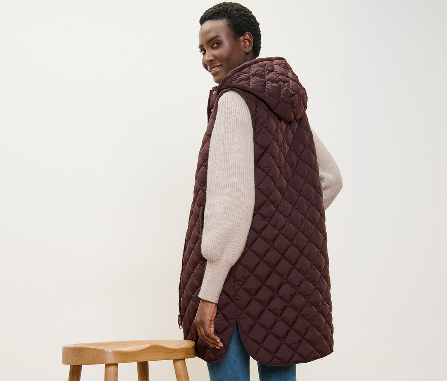 Žena se usmívá přes rameno a má na sobě dlouhou prošívanou vestu s kapucí.