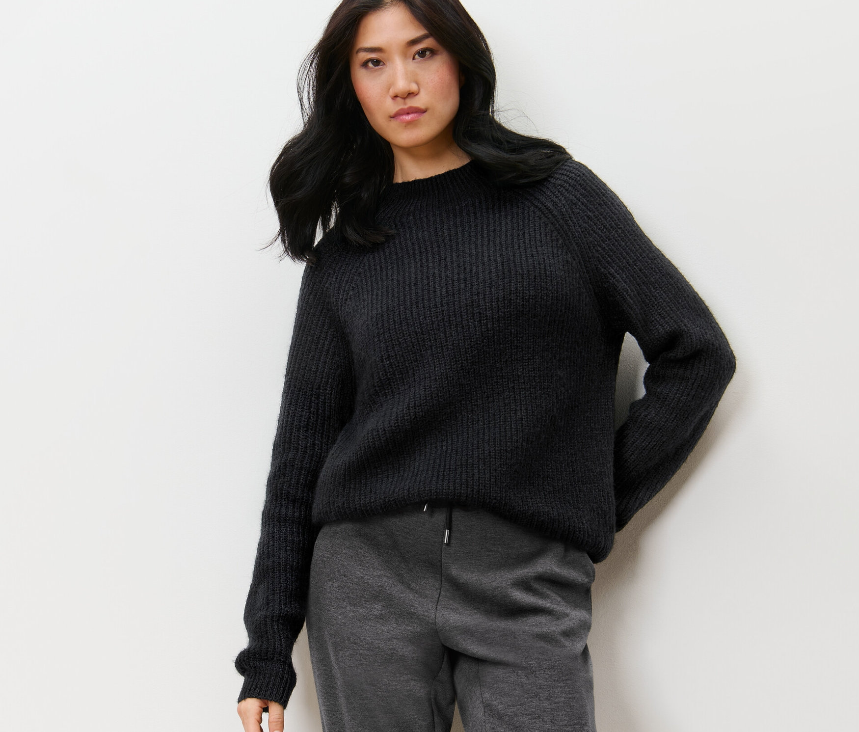 Žena pózuje na pozadí stěny a má na sobě černý svetr z hrubé pleteniny se stojáčkem.