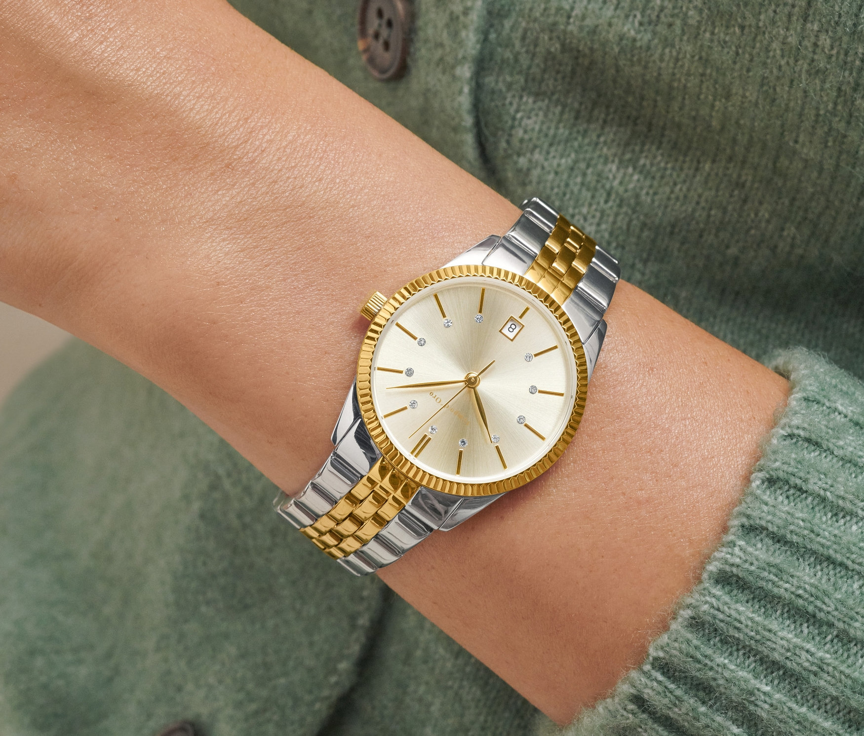 Detail dámských hodinek ve stříbrno-zlaté kombinaci s datumem na zápěstí ženy v zeleném svetru.